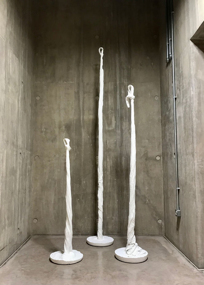 Three white rope like sculptures in a grey cement walled space.
