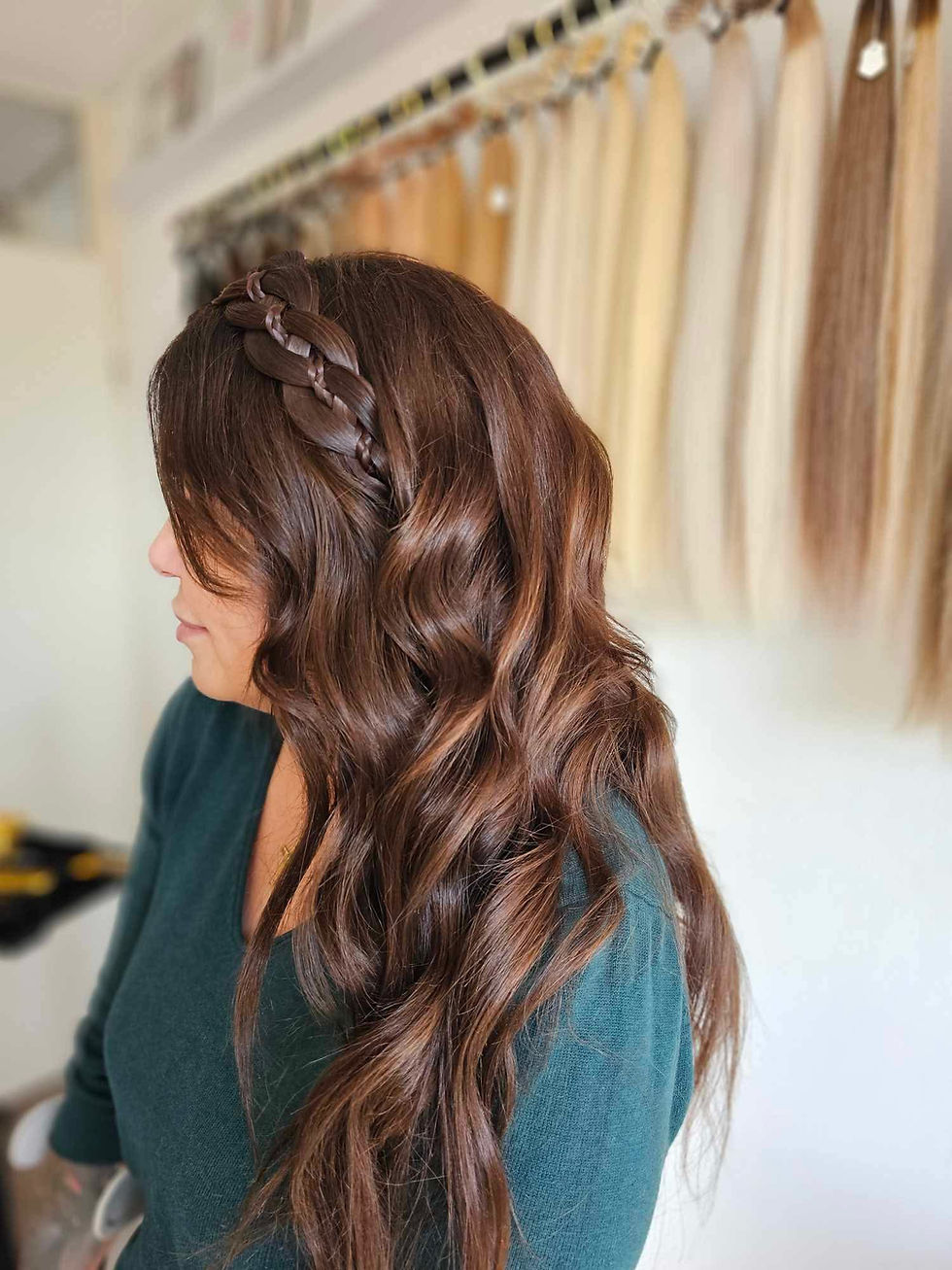 Woman with wavy brown hair extensions and braid stands in front of hair extensions display. Wears a teal sweater