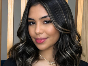 Mulher de cabelo preto ondulado com mechas, sorrindo levemente, usando colar de infinito, diante de painel de madeira.