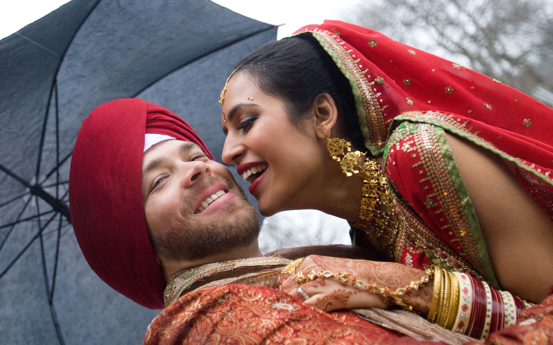 A mixed race couple as part of a larger Sikh wedding gather under their umbrella during a January winter wedding © 2024 Michael Buescher Wedding Photography #bestvancouverweddingphotographers