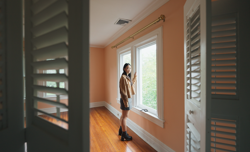 Woman in a brown jacket stands by a window in an empty room with peach walls and wooden floors. She looks outside, creating a serene mood.