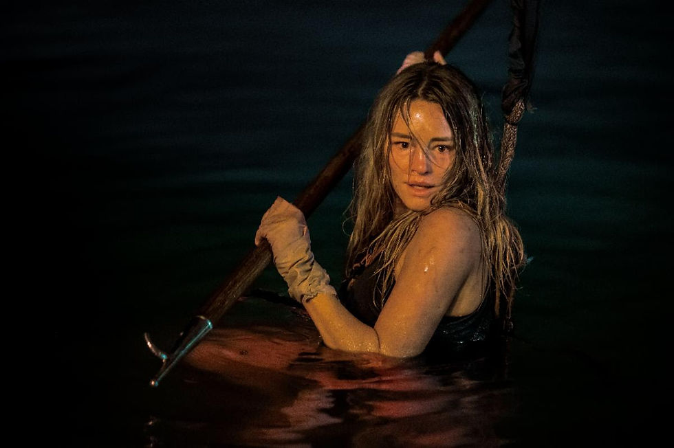 A woman in water, gripping a pole, looks intensely toward the camera. Dark, moody background with greenish hues.
