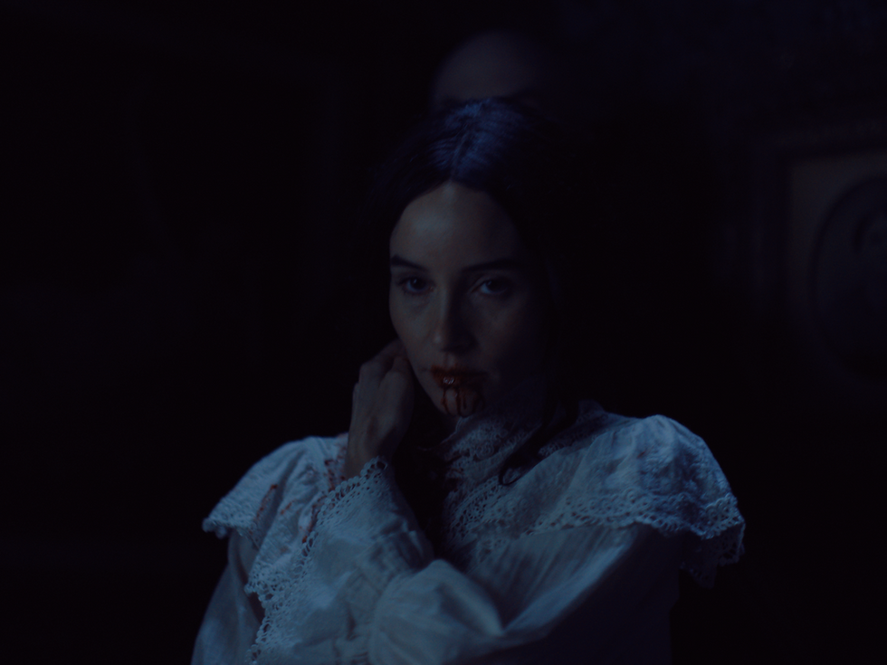 Woman with blood on her mouth wears a white lace dress in a dark, eerie setting. The mood is mysterious and haunting.