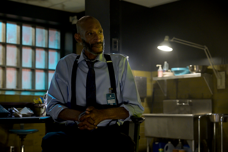 Man in a shirt and tie, seated in a dimly lit room, illuminated by a desk lamp. Background includes a window and kitchen sink. Quiet mood.