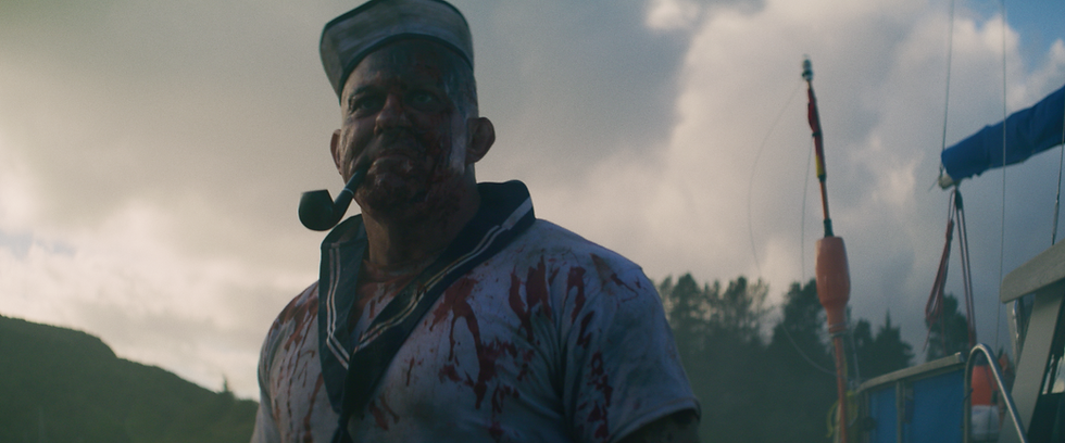 Bloodied sailor with a pipe stands on a boat, wearing a stained uniform against a cloudy sky and trees, creating a tense mood.