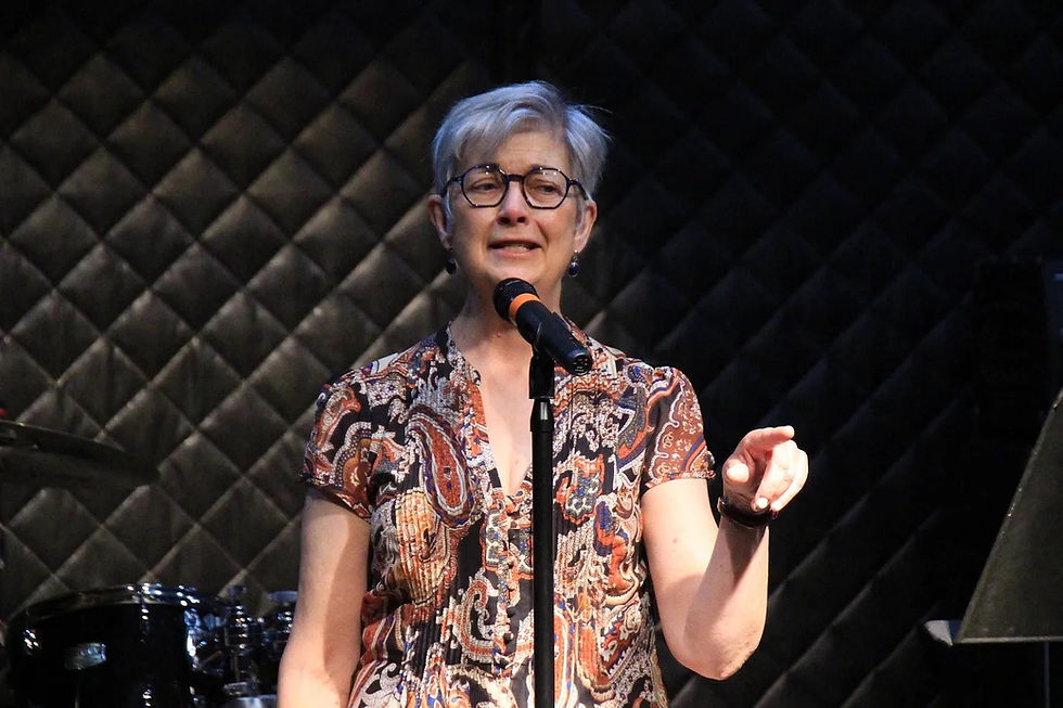 Woman with glasses singing into a microphone on stage, wearing a colorful patterned shirt. Quilted background, drum set nearby.
