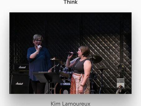 Two women sing into microphones on a small stage, with drum kit and Marshall and Yamaha gear against a black quilted backdrop.