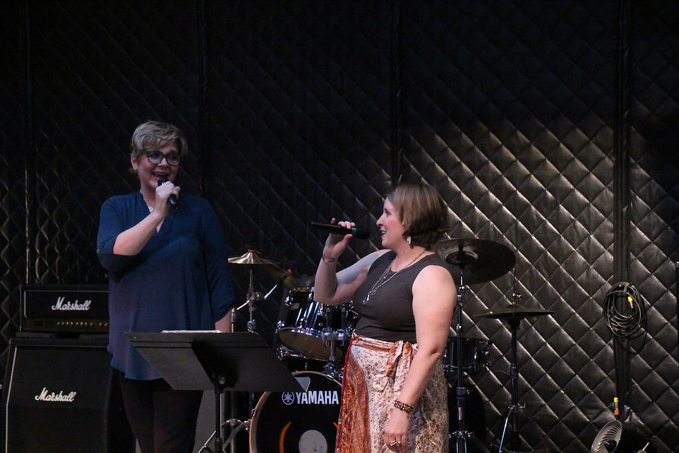 Two women sing into microphones on a small stage, with drum kit and Marshall and Yamaha gear against a black quilted backdrop.