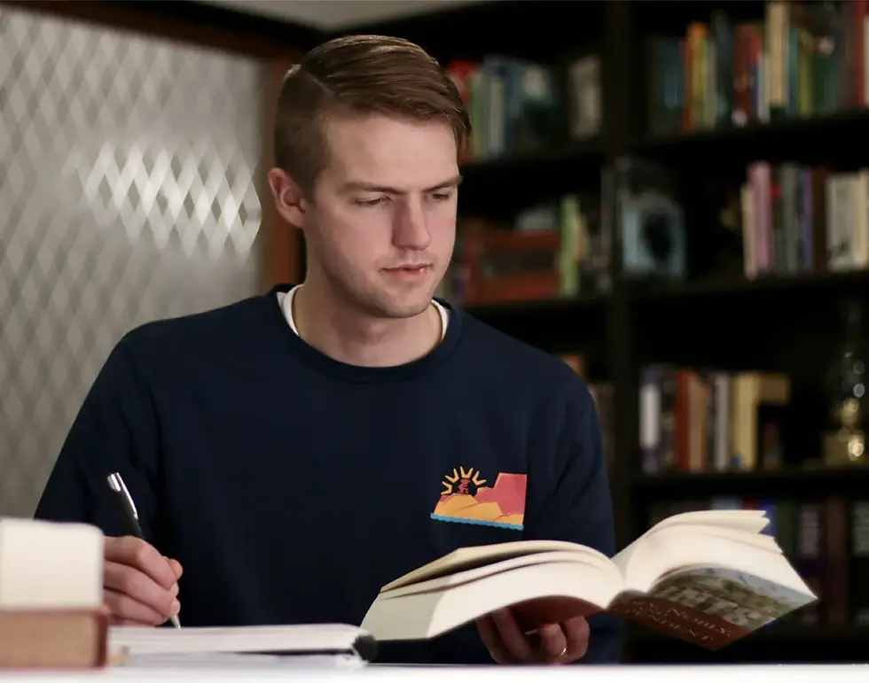 A student in an NDP shirt reads and takes notes at a desk in front of a bookshelf