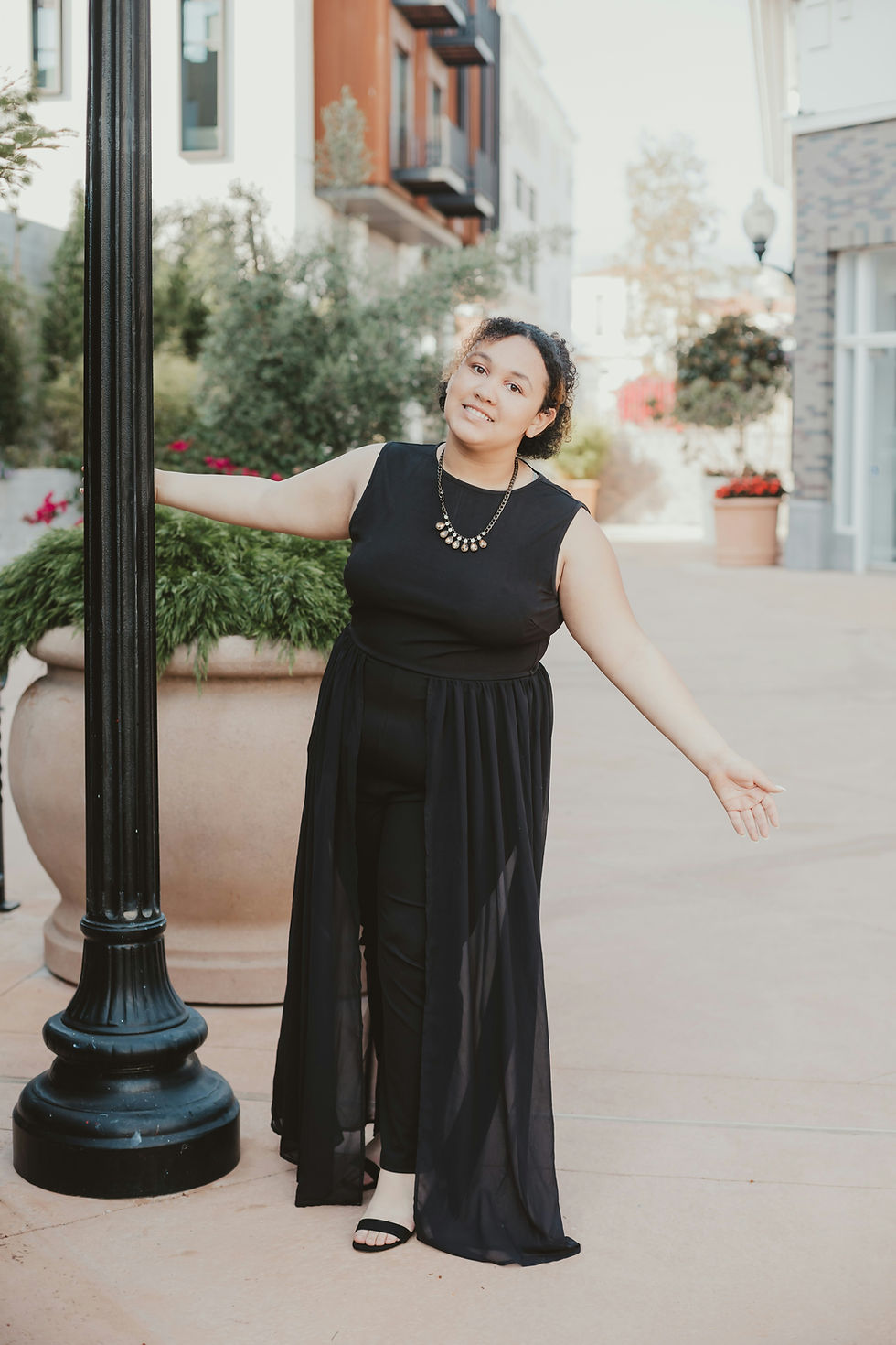 Woman in black dress and necklace poses joyfully by a lamppost in an outdoor setting with buildings, greenery, and warm lighting.