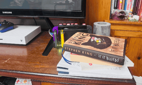 Stephen King's "If It Bleeds" book sits on papers beside a gaming console and TV. The desk is cluttered, creating a cozy, busy vibe. The animation shows the stack transformed from just a stack of papers and a book to a stack that has been corralled into a nice blue cotton basket.