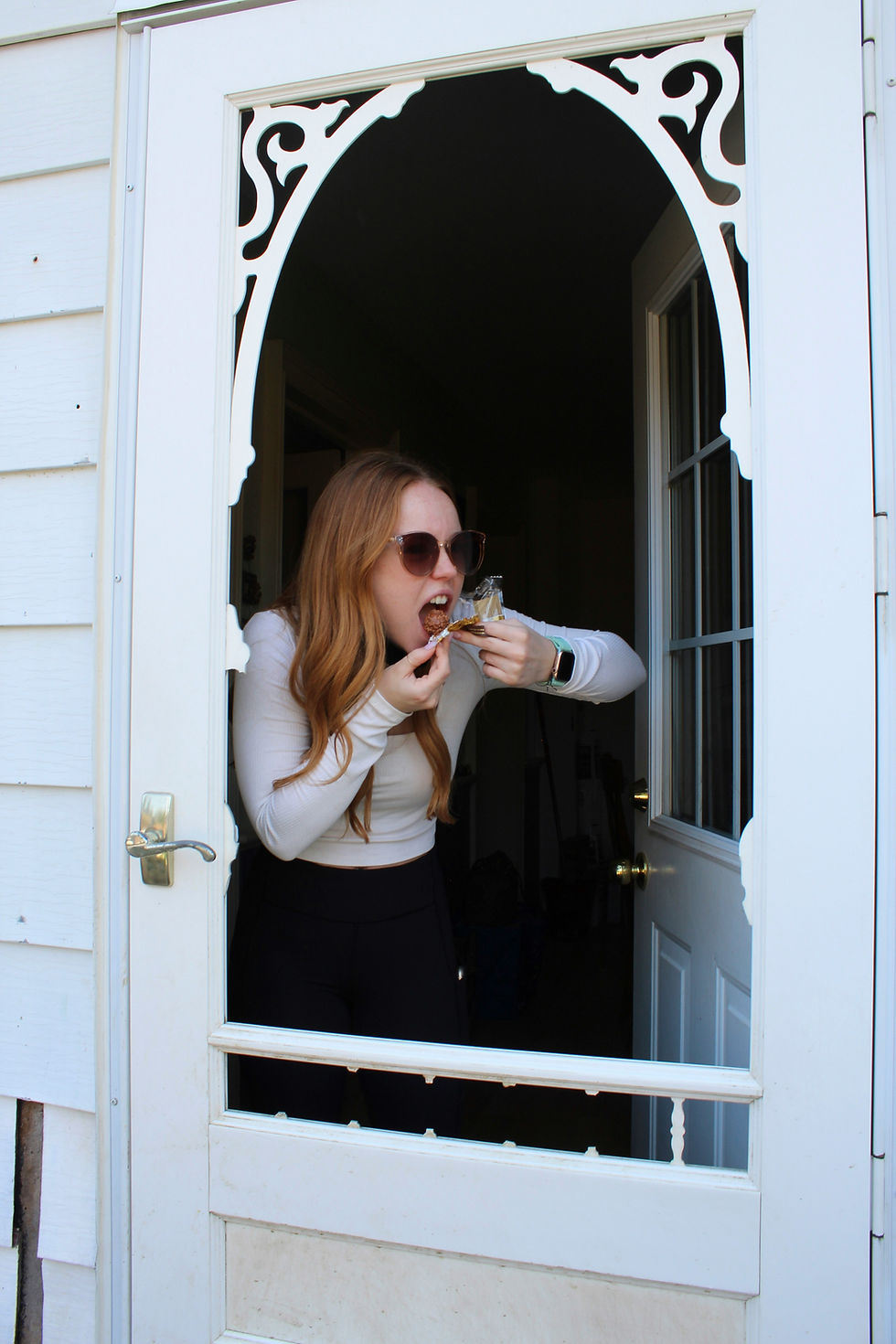 Woman in sunglasses eats a piece of chocolate at decorated white door. She is wearing a white long-sleeve top, black pants, and a smartwatch.