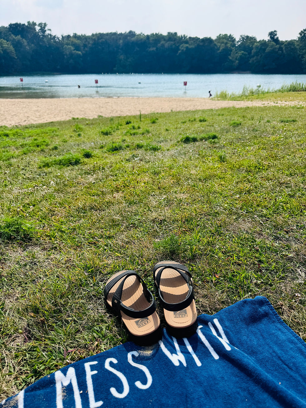 Sandals on a blue towel with "MESS WITH" text on grass. Sandy beach, calm lake, and wooded background under a clear sky. Relaxing mood.