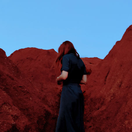 A women in a long black dress with back turned, and looking downward amongst red rocks and a cloudless dim blue sky.