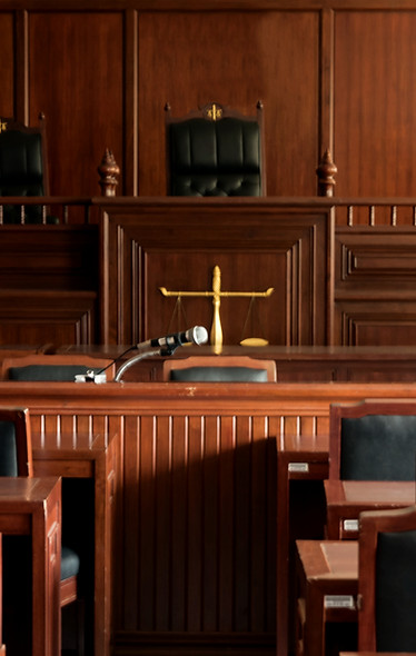 An empty wooden courtroom featuring wooden benches, a microphone on the central table, and scales of justice in front of the judge's bench.