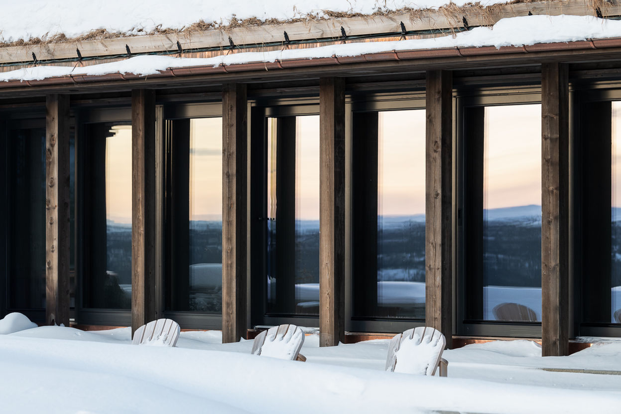 Grånede tresøyler foran store vindusfelt og skyvedører med panorama utsikt mot fjellheimen. Svalgangs motiv med dører til skifer stein terrassen.