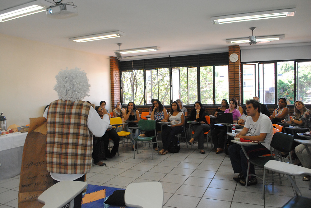 apresentação do monólogo. A personagem está de pé, usa peruca da cor branca, camisa de manga longa branca também e um colete xadrez. Os participantes estão sentados, observando-a.