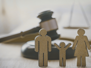 A wooden figure of a family; parents and child, standing together in front of a gavel and law scale.