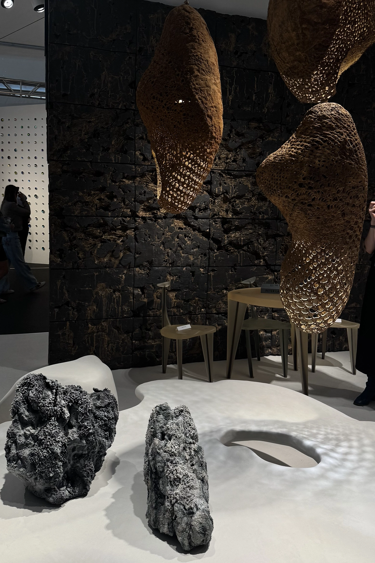 Sculptural collectible design installation at Design Miami featuring biomorphic hanging forms, dark textured walls, mineral-like stone elements and a white organic floor surface, highlighting raw materiality and tactile interior trends.