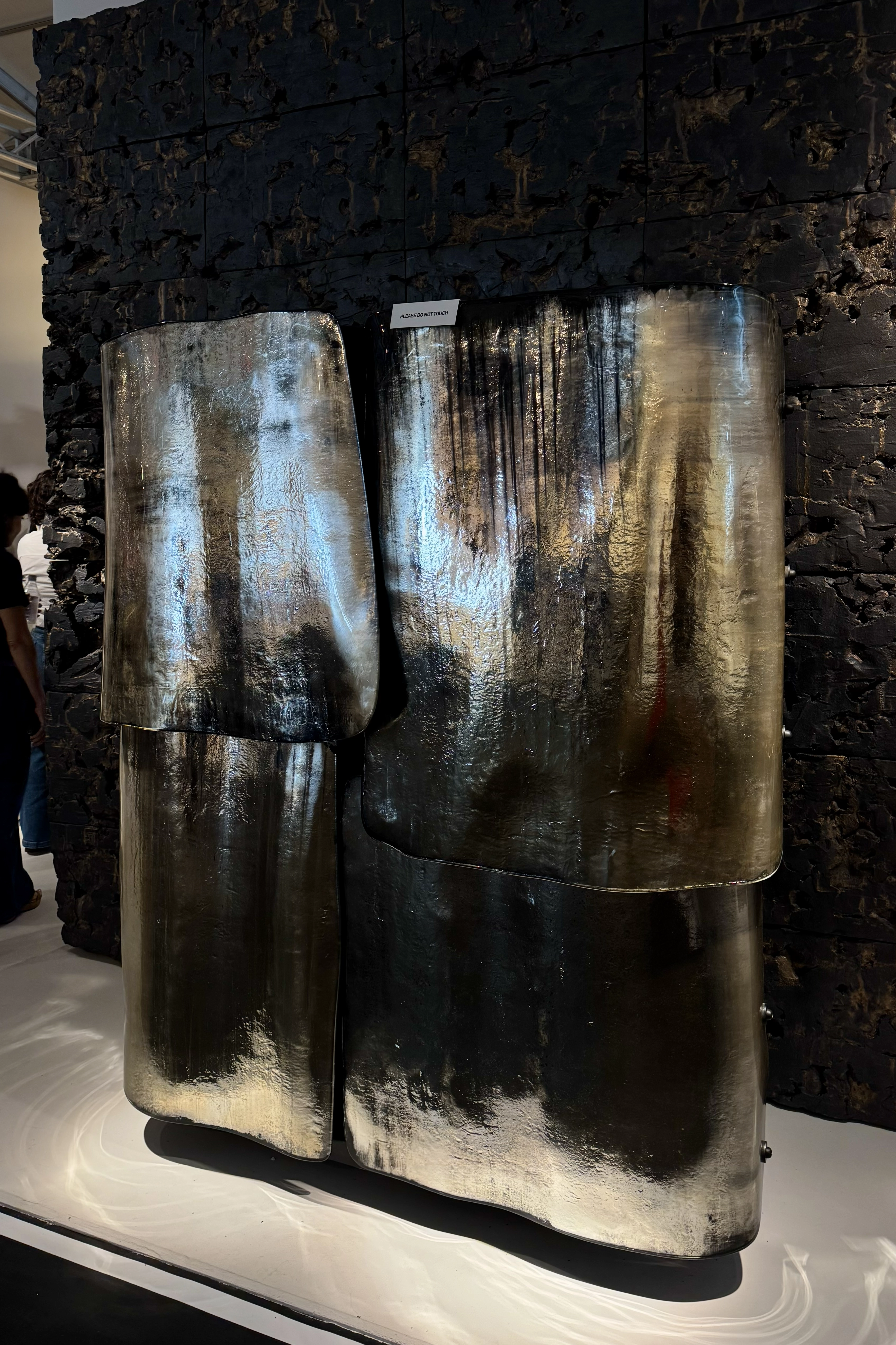 Collectible design installation featuring sculptural metal panels with raw, oxidized textures and reflective surfaces, displayed against a dark textured wall in a contemporary exhibition space