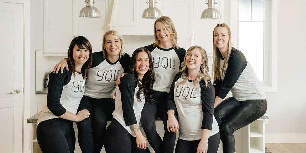 Six women in matching "YQL" shirts pose happily in a modern kitchen, with white cabinets and silver pendant lights, creating a cheerful mood.