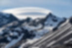 Snow-covered mountains with a unique cloud formation above, under a clear blue sky, creating a serene and majestic landscape.