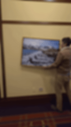 Person steadying a tilted penguin MetalPrint on a beige wall. The frame has a gold border. Carpet with red and brown patterns below.
