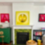 A room with colorful framed artwork on white walls, a green-tiled fireplace, and shelves of books. Bright and artistic setting.
