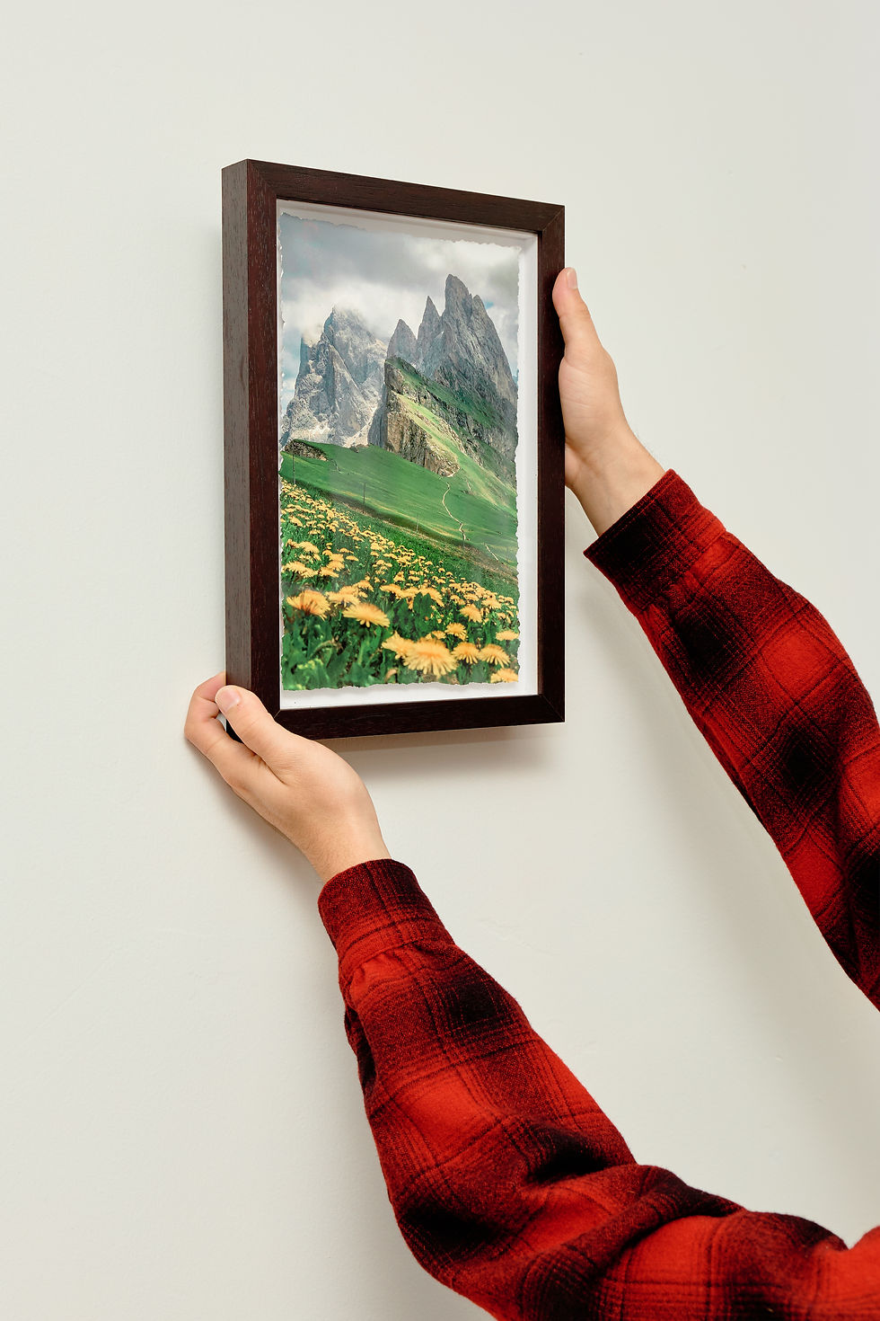 Hands in red plaid sleeves hang a Framed Print with green hills and yellow flowers on a white wall.