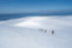 Four people in bright jackets hike on a snowy slope towards the sea. A ship and rock formations are visible under a clear blue sky.