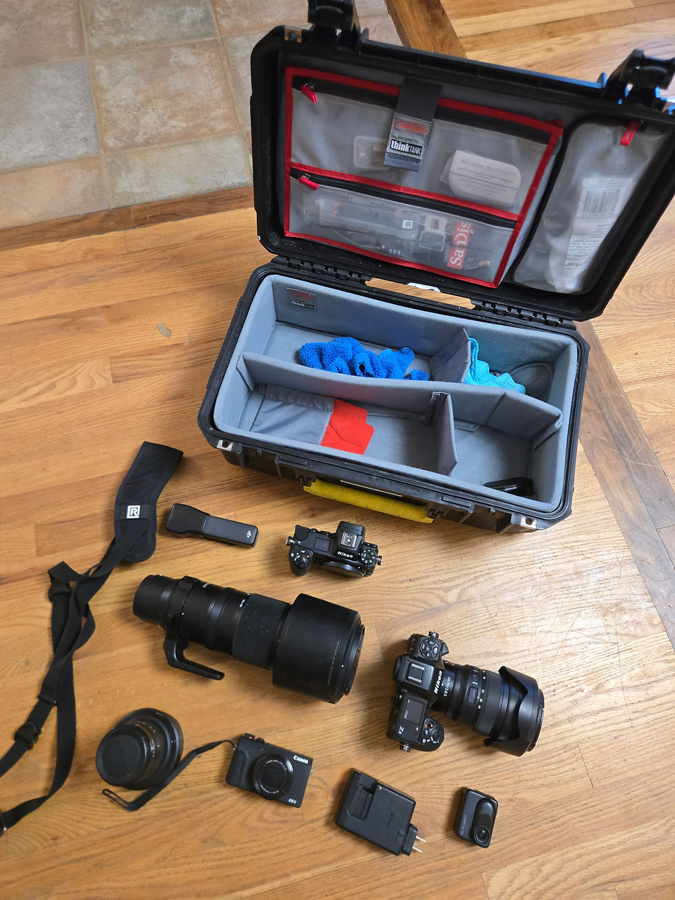 Camera gear on wooden floor: open black case, lenses, cameras, and straps. Visible "Think Tank" and "SanDisk" text. Modern photography setup.