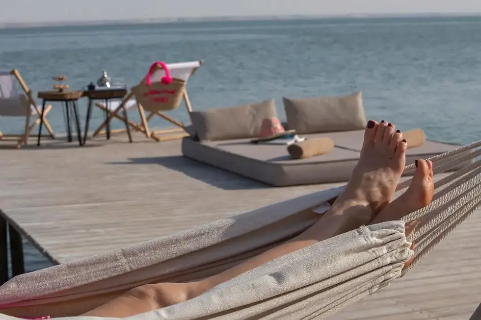 Pieds nus dans un hamac sur une terrasse au bord de l’eau, avec transats et mer calme au fond.