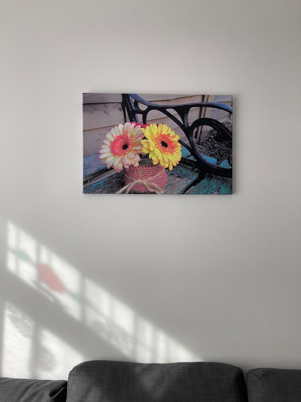 Thumbnail: A canvas print on a white wall featuring a photograph of gerberas on an old bench. Grey pillows are visible at the bottom of the image.