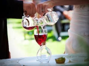 bride and groom pour small jugs of red wine into a large wine glass for a wine ceremony