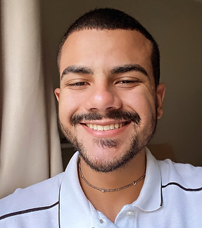 A imagem é de um jovem homem sorrindo. Ele tem cabelos curtos e escuros, com um cavanhaque e bigode que acompanham o contorno de sua boca. Ele usa um colar fino com pequenas contas prateadas. Sua camiseta polo é branca com detalhes em preto nas golas e nas mangas. A iluminação sugere que ele está perto de uma janela, com a luz vindo de um lado. O fundo está levemente desfocado, com uma cortina visível.
