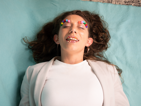 Alex lays down with pom poms on eye brows and sprinkles on lips with eyes closed