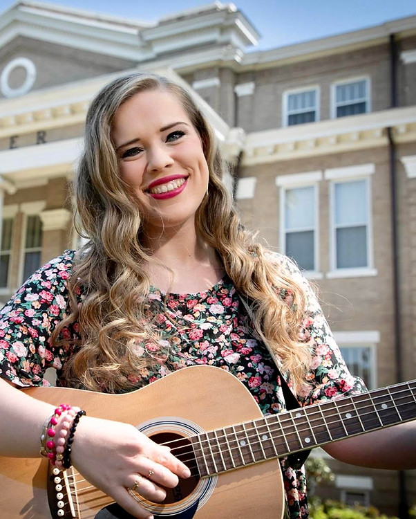 Caucasian woman in her 20s with long curly hair, playing a guitar in front of a building.