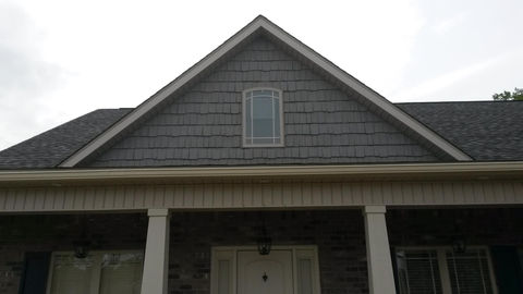 Front view of a house with a steep gable roof, gray shingle siding on the upper facade, a small arched window, and a covered porch supported by white columns.