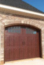 Dark wooden garage door with decorative windows and handles, framed by an arched brick entryway on a brick house exterior.