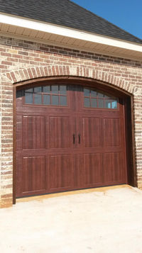 Brown garage door with decorative windows and an arched brick entryway, set against a brick exterior wall.