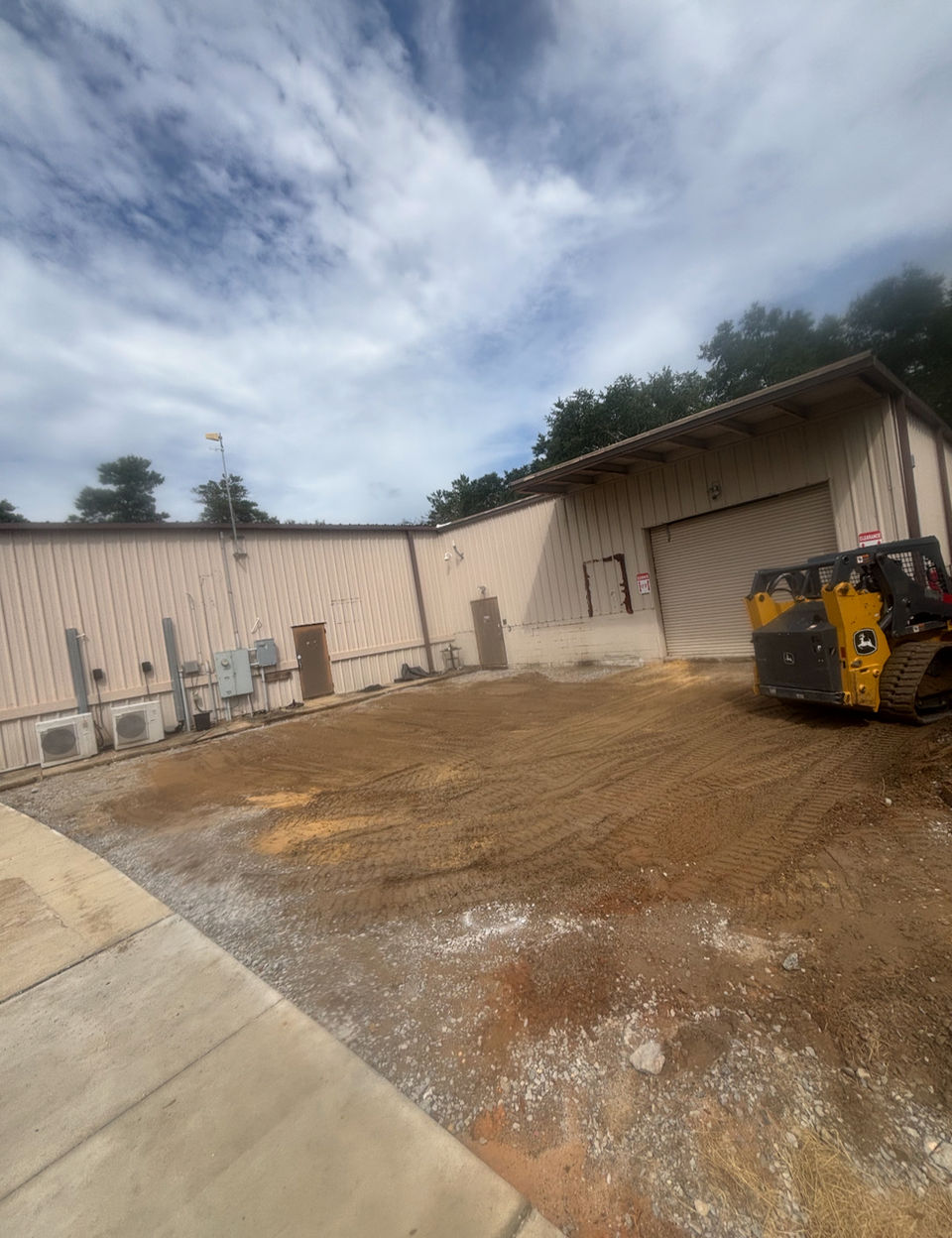 Construction site beside a beige industrial building where a skid steer loader is leveling dirt for grading or foundation preparation under a partly cloudy sky.