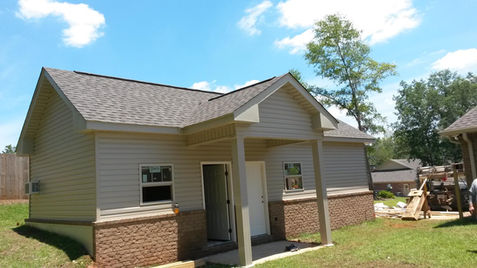 Compact single-story home with tan siding, brick foundation, covered front porch, and a grassy yard under a sunny blue sky.