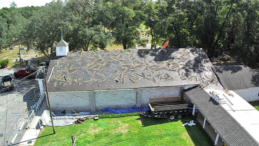 Aerial view of a large building roof under renovation with workers removing old materials, surrounded by trees and equipment, including a trailer parked on the grass below.