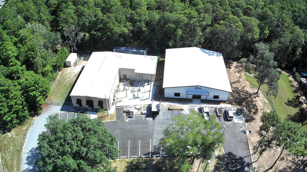 Aerial view of a commercial construction site with two large metal buildings surrounded by trees, featuring paved parking areas and ongoing exterior work.