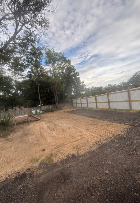 Leveled dirt lot prepared for construction or parking, bordered by a wooden fence and surrounded by trees under a partly cloudy sky.