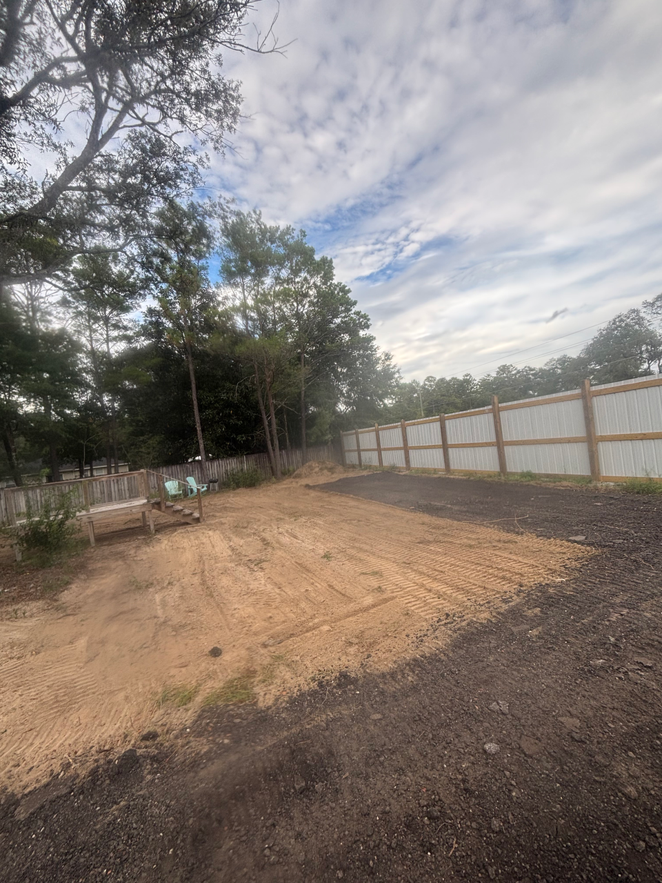 Leveled dirt lot prepared for construction or parking, bordered by a wooden fence and surrounded by trees under a partly cloudy sky.