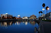 Waterfront cityscape at dusk featuring buildings, palm trees, and reflections of lights on the calm water.