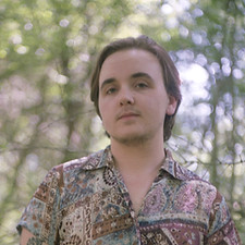 portrait photo de Riv Espaignet, personne transmasc, il regarde la caméra avec fierté, il a les cheveux courts, il porte une chemise à motifs