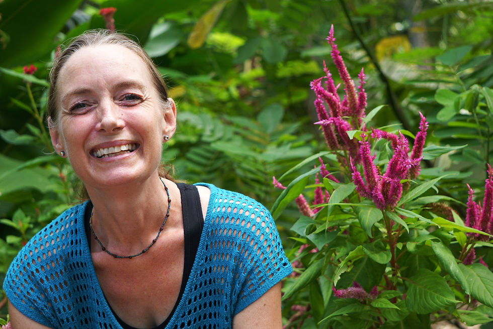 Nat smiling a in blue top, sitting by vibrant pink flowers, surrounded by lush green foliage in a garden setting.