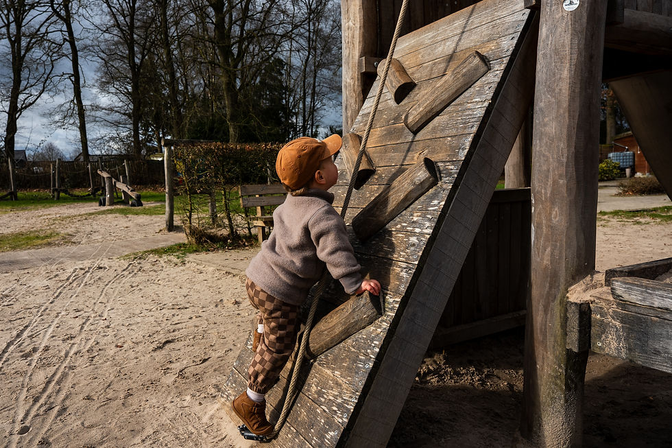 Kind speelt buiten op houten klimrek met touw, draagt bruine pet en geruite broek. Zonnige dag, bomen en zand in achtergrond.